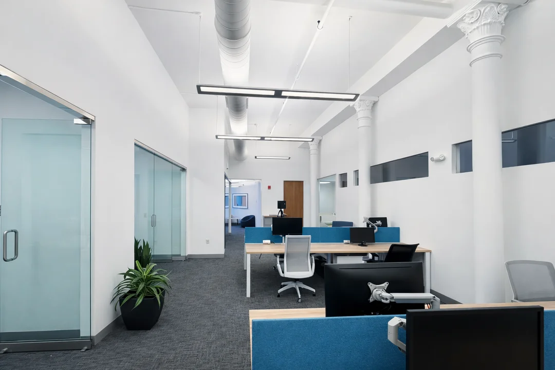 Contemporary open office interior featuring shared workstations, glass-walled offices, exposed ductwork, and tall columns.