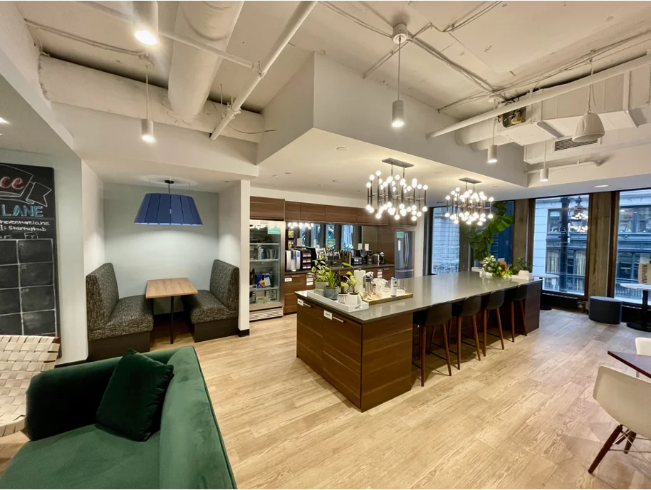 Contemporary office breakroom featuring a wood-finished kitchen island with bar stools, chandelier lighting, built-in appliances, and cozy booth seating.