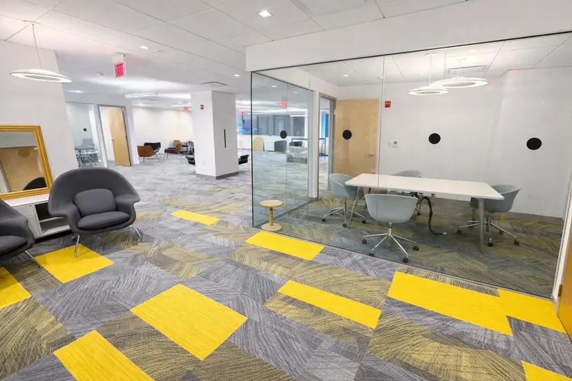 Contemporary office space featuring a glass-walled conference room, casual lounge chairs, and gray carpet with yellow accent tiles.