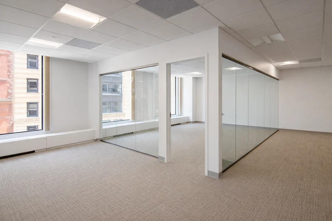 Bright, vacant office suite featuring floor-to-ceiling windows, glass partitioned meeting rooms, white walls, and beige carpet flooring.