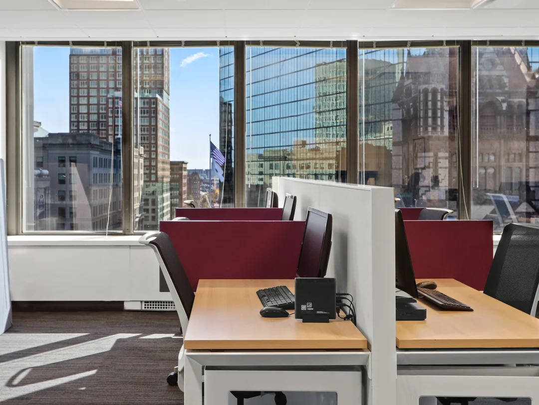 Modern office workspace with two desks and computer monitors facing large windows overlooking a downtown city skyline.