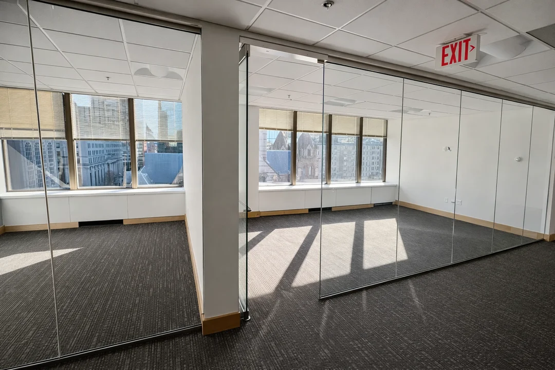 Vacant office interior featuring floor-to-ceiling mirrored glass partitions, dark carpet flooring, and large windows overlooking downtown buildings.