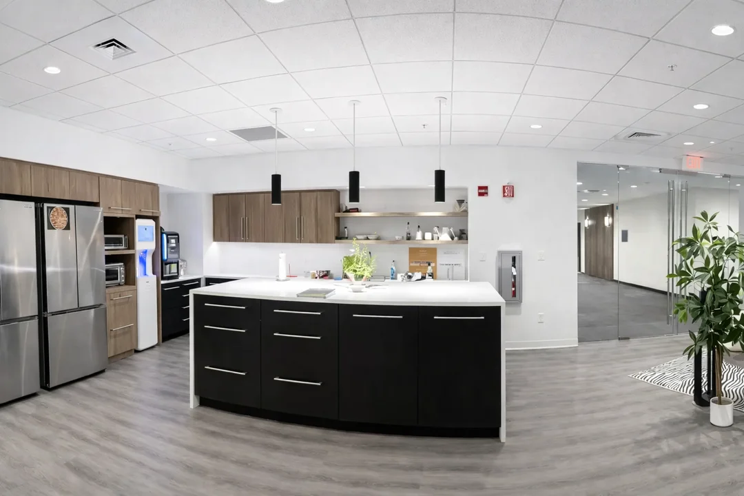 Contemporary workplace kitchen featuring a large island with pendant lighting, stainless steel refrigerator, coffee station, and open shelving.