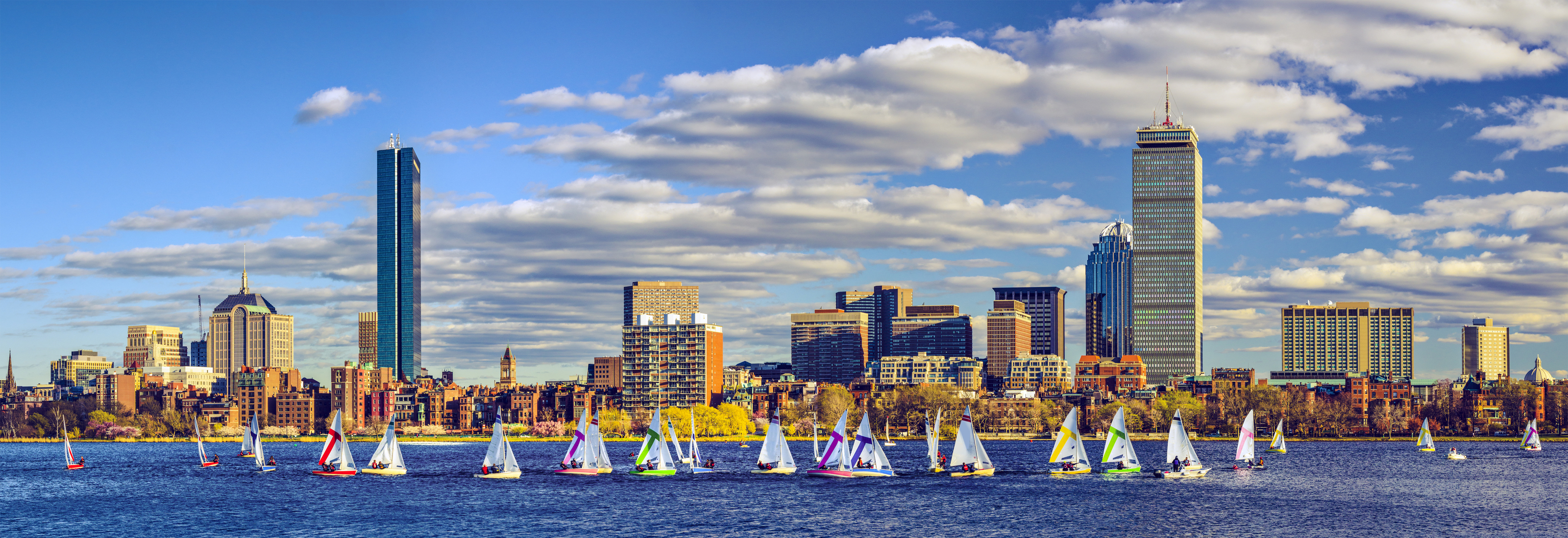 Panoramic view of the Boston skyline, including modern skyscrapers and historic buildings, with colorful sailboats on the Charles River.