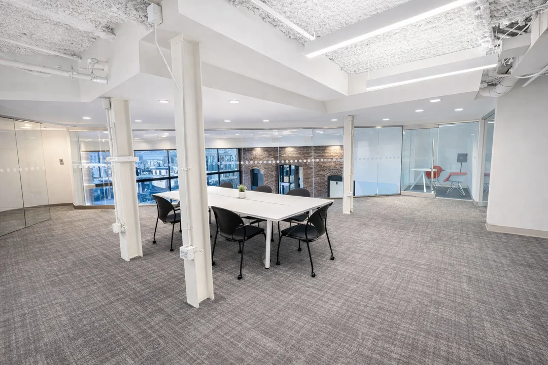 Modern office interior featuring a central collaboration table, glass conference rooms, exposed beams, and large windows with brick accents.